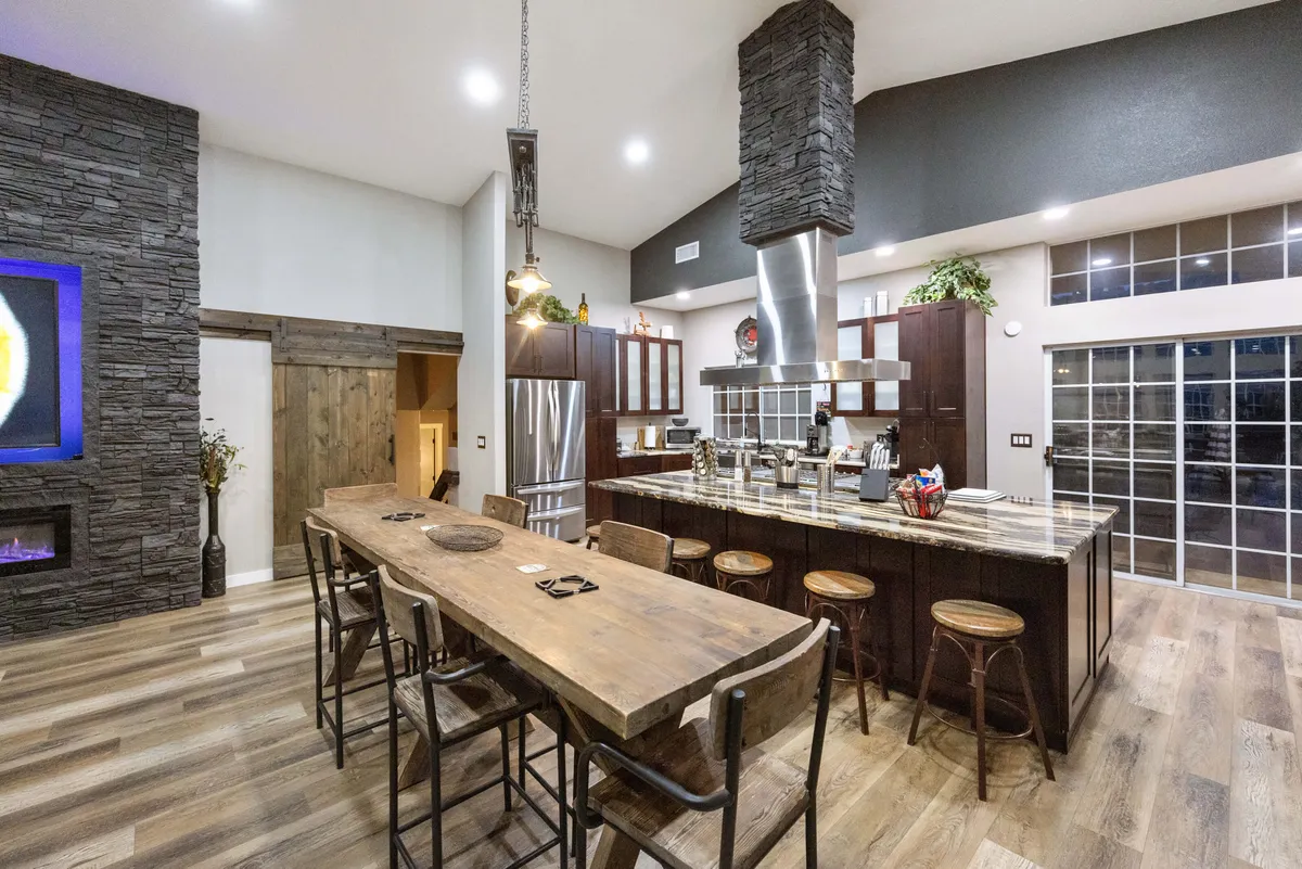 A spacious open-plan kitchen and dining area, featuring a large wooden dining table ideal for group meals, a kitchen island for prep and conversation, stainless steel appliances, and a striking stone feature wall. The atmosphere is communal, inviting, and perfect for celebrations.