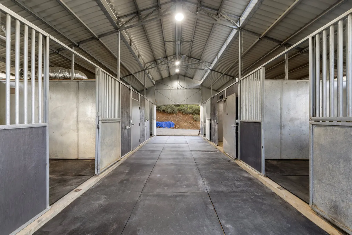 A bright indoor horse stable with neatly organized stalls, an aisle, and warm lighting. The area looks clean and spacious, emphasizing high-quality equestrian facilities for horse lovers.