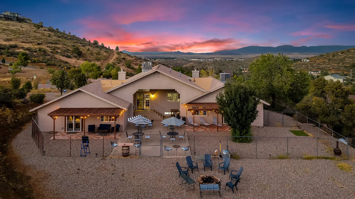 This exterior shot highlights the house with its inviting patio, a central fire pit area, and a stunning mountainous backdrop beneath a colorful sunset sky. The scene is set for outdoor gatherings, storytelling by the fire, and relaxing while taking in the hillside views, all key to the team-building and relaxation theme.