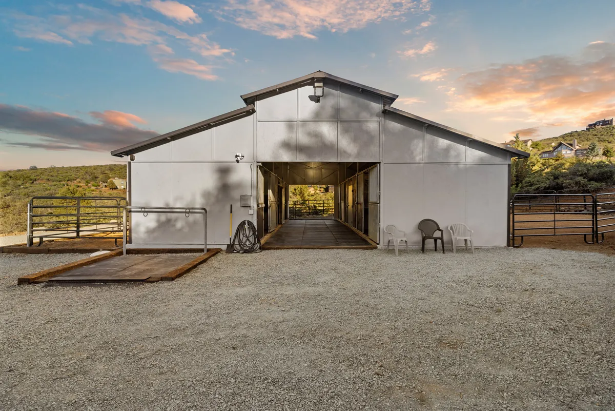 A tranquil exterior view featuring a metal barn structure with a pathway leading inside, surrounded by natural landscape and outdoor chairs. The scene is bathed in soft light, with hints of sunset visible on the horizon. The barn's location and setup evoke an inviting environment for equestrian activities and direct access to the wilderness.