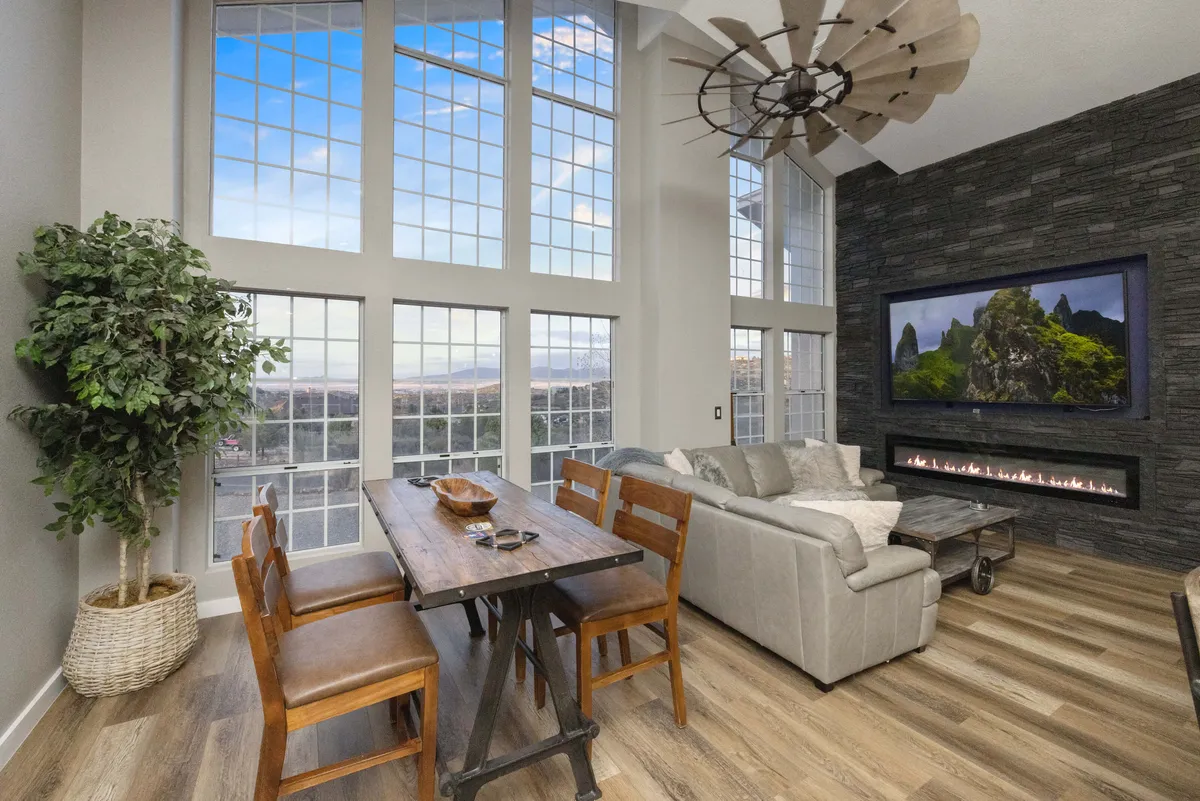 A cozy, modern living room with large windows offering sunset views, a dining table for shared meals, a sofa for relaxing, a fireplace, and a TV. The setting is designed for family gatherings, dining, and unwinding together.