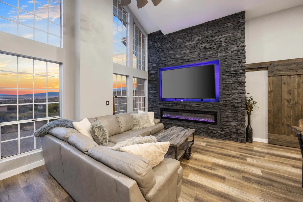 A spacious living area featuring large windows that reveal scenic hillside views, a comfortable sofa set, a stylish stone feature wall, a TV, and a welcoming fireplace. The décor balances elegance and rustic charm, inviting guests to relax, socialize, or enjoy the views.